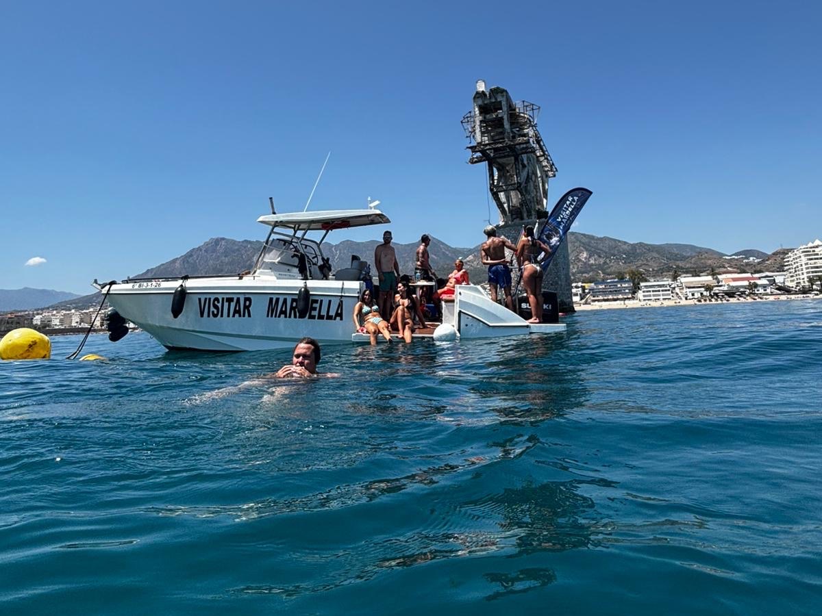 The Visitar Marbella dive boat at anchor beside the crane structure of The Tower dive site off Marbella, with swimmers in the water and La Concha mountain behind