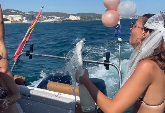 The bride spraying champagne at the stern of the Visitar Marbella boat, balloons and the coast behind