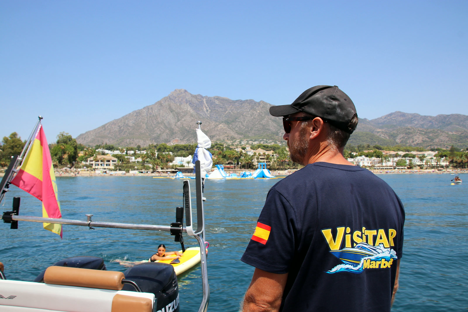 Jason at the helm in a Visitar Marbella shirt, watching a guest on the paddleboard, La Concha and the Marbella coast behind