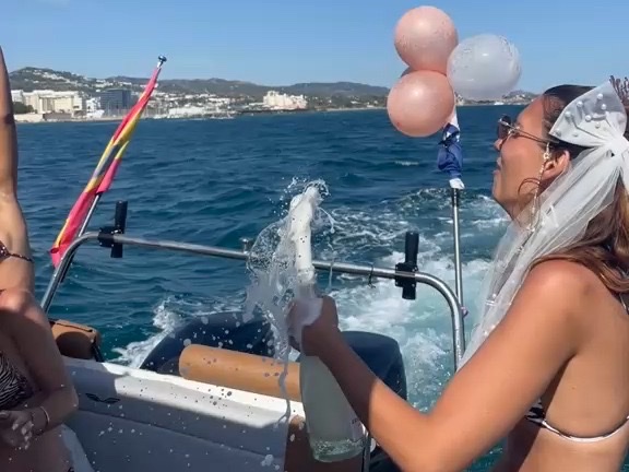 The bride popping prosecco aboard the Visitar Marbella boat, veil and balloons flying, the Marbella coastline behind